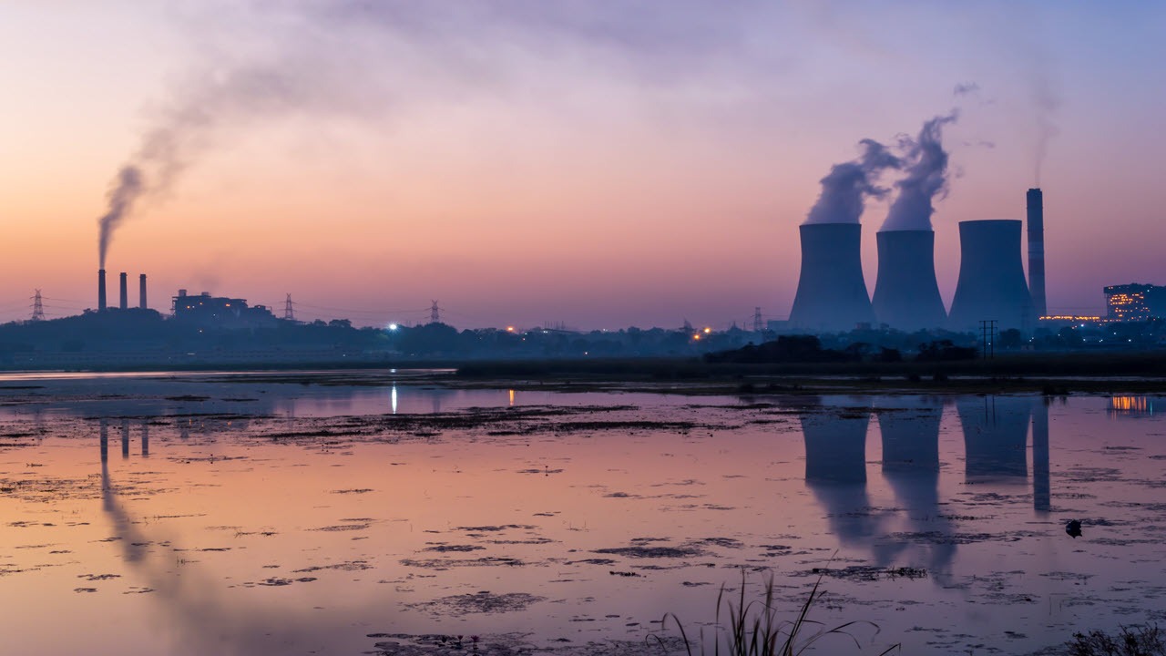Industrial power plant with cooling towers and smokestacks releasing emissions at dusk, reflected across a water body, illustrating environmental impact and water pollution concerns.