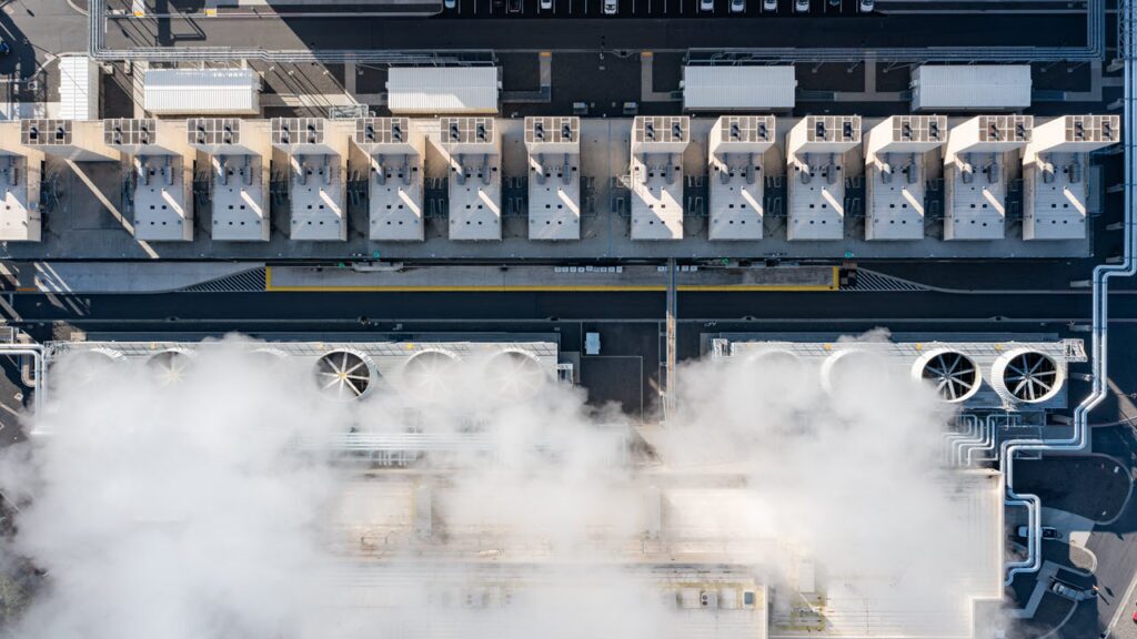 Aerial view of data center cooling systems with rooftop units, large fans, and vapor plumes, illustrating infrastructure required for reliable thermal management in hyperscale AI and cloud facilities.